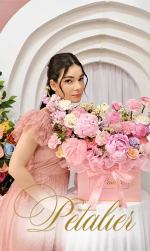 A woman standing and leaning on a table with her hand beside a box of mixed flowers, with the word ‘Petalier’ in stylized font